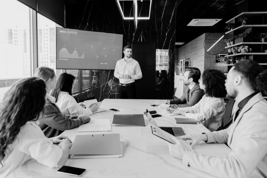 Business meeting with a man presenting annual statistics on a screen to a group of colleagues seated around a conference table.