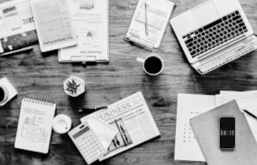 Overhead view of a cluttered workspace with a laptop, newspapers, notebooks, coffee cups, a smartphone, and various documents.