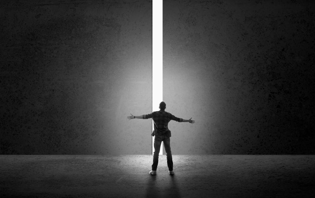 Man standing in front of a tall wall with a narrow opening of light, arms wide open, symbolizing leadership evolution and embracing challenges.